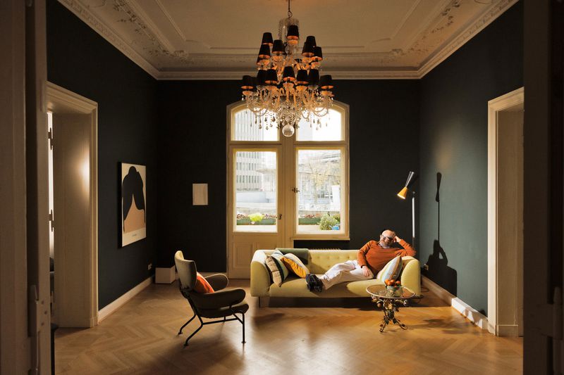 A man in an orange sweater reclines on a sofa in a dark-walled living room beneath an ornate chandelier with parquet flooring.
