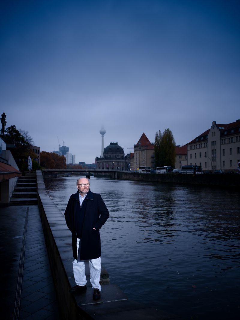 Man in a dark overcoat and white trousers standing on a riverbank promenade in Berlin at dusk with city landmarks behind him.