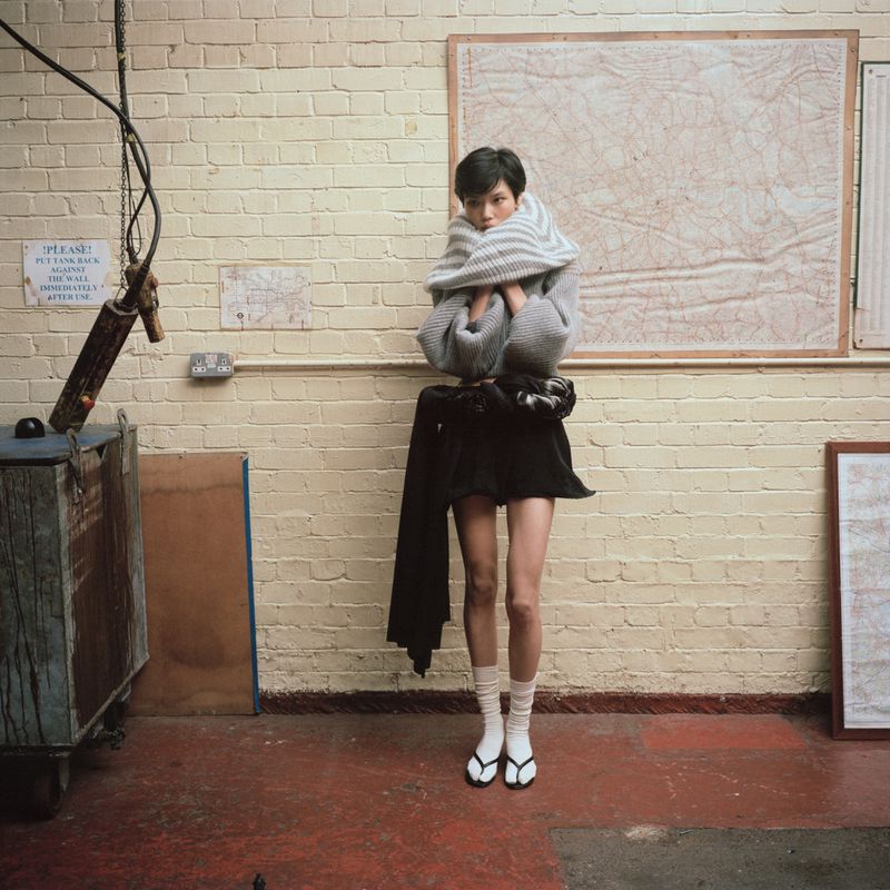 Model standing against a painted brick wall wearing an oversized grey knit wrap, black miniskirt, white socks, and black sandals.