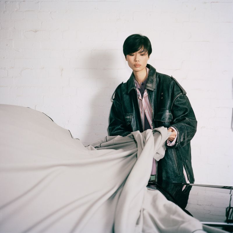 An East Asian model in an oversized black leather jacket lifting a light grey fabric cover from a car against a white wall.