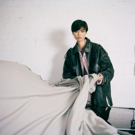 An East Asian model in an oversized black leather jacket lifting a light grey fabric cover from a car against a white wall.