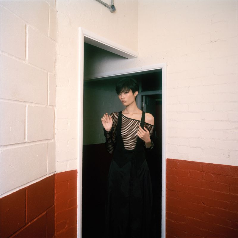 Model with short black hair standing in a doorway wearing a black mesh top and skirt against a painted brick wall
