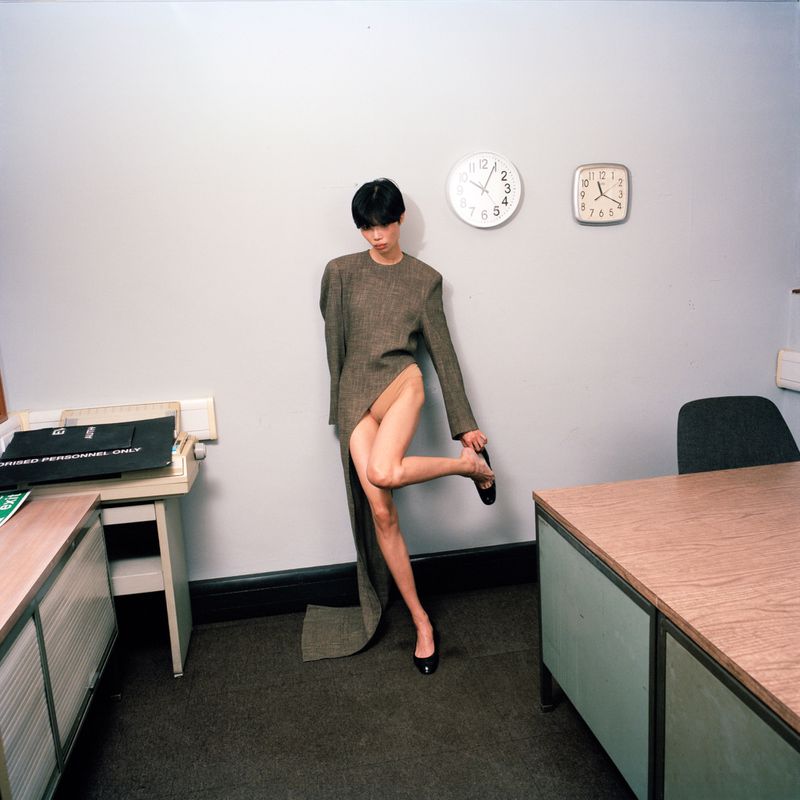 Model wearing asymmetric brown dress standing in office with desks and two wall clocks for Harper's Bazaar France KAYAKO