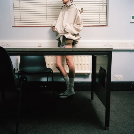 Model wearing an oversized beige hooded jacket and grey socks standing behind an office desk in front of closed blinds.