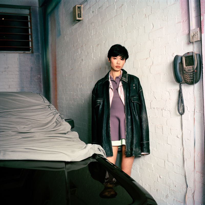 Woman in dark leather jacket standing against a white brick wall beside a covered car and wall phone