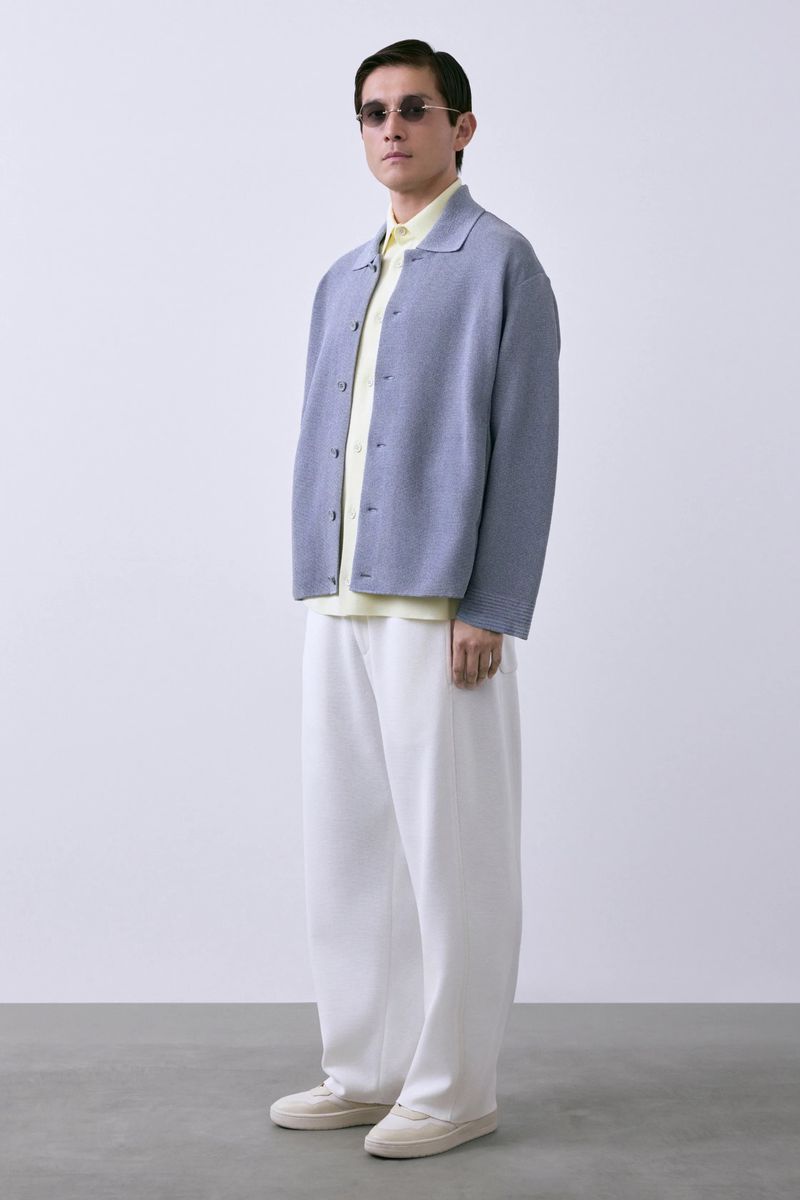 Man in a light blue knit jacket, yellow shirt, and wide-leg white trousers standing against a pale studio background.