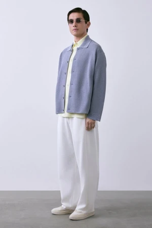 Man in a light blue knit jacket, yellow shirt, and wide-leg white trousers standing against a pale studio background.