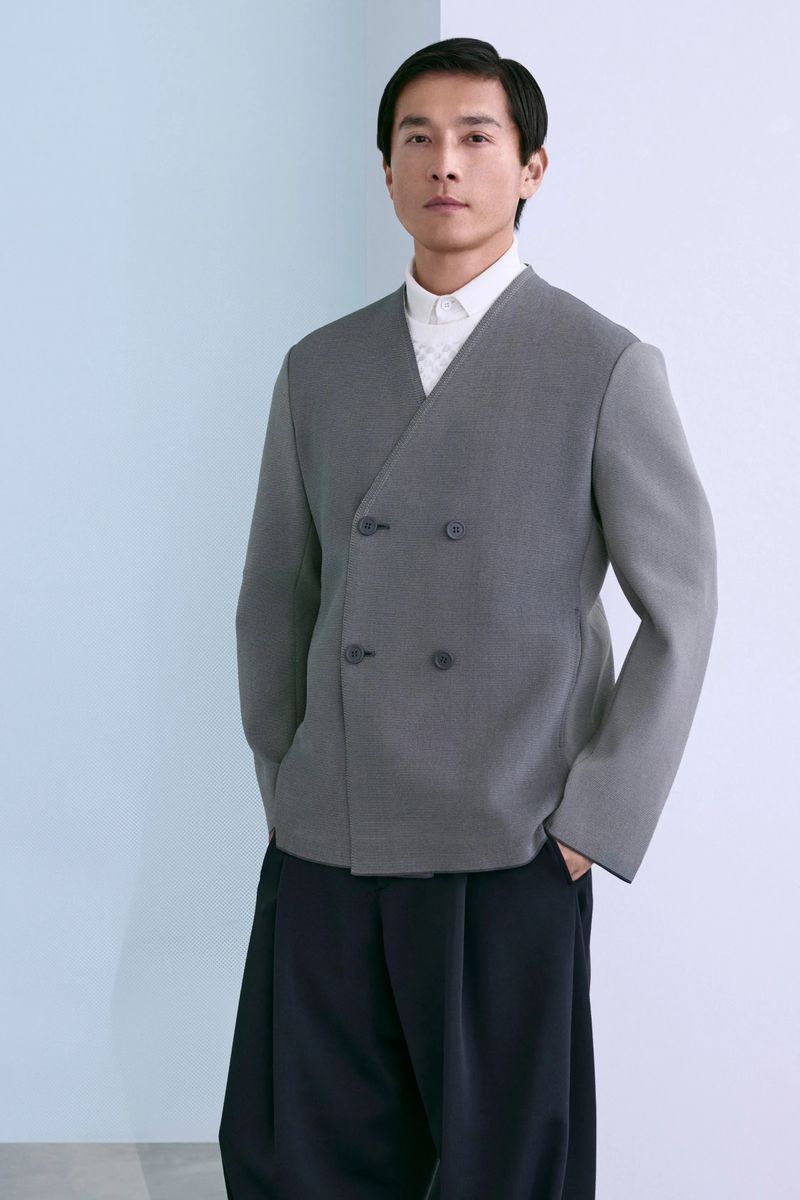 Model Daisuke Ueda in a grey double-breasted collarless knit jacket and dark wide-leg trousers against a pale studio backdrop.