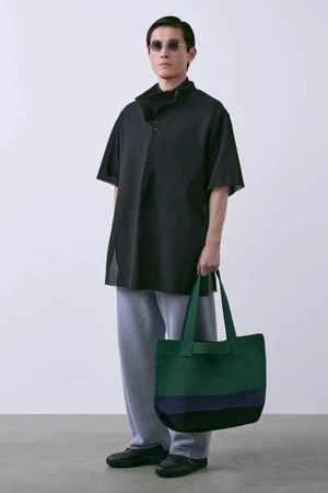 Model wearing sunglasses, a black short-sleeved tunic shirt, grey knit trousers, and holding a green tote bag against a neutral background.