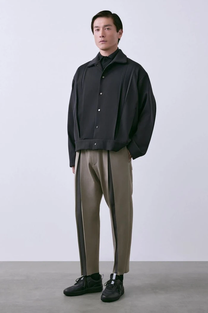 Model Daisuke Ueda stands in a black structured snap-button jacket and taupe trousers against a pale studio backdrop for CFCL.