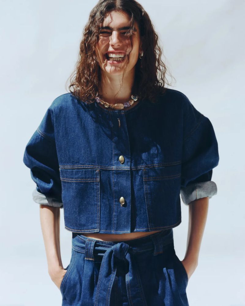 Julia Pacha laughing in a cropped dark blue denim jacket and belted denim trousers against a white studio background.