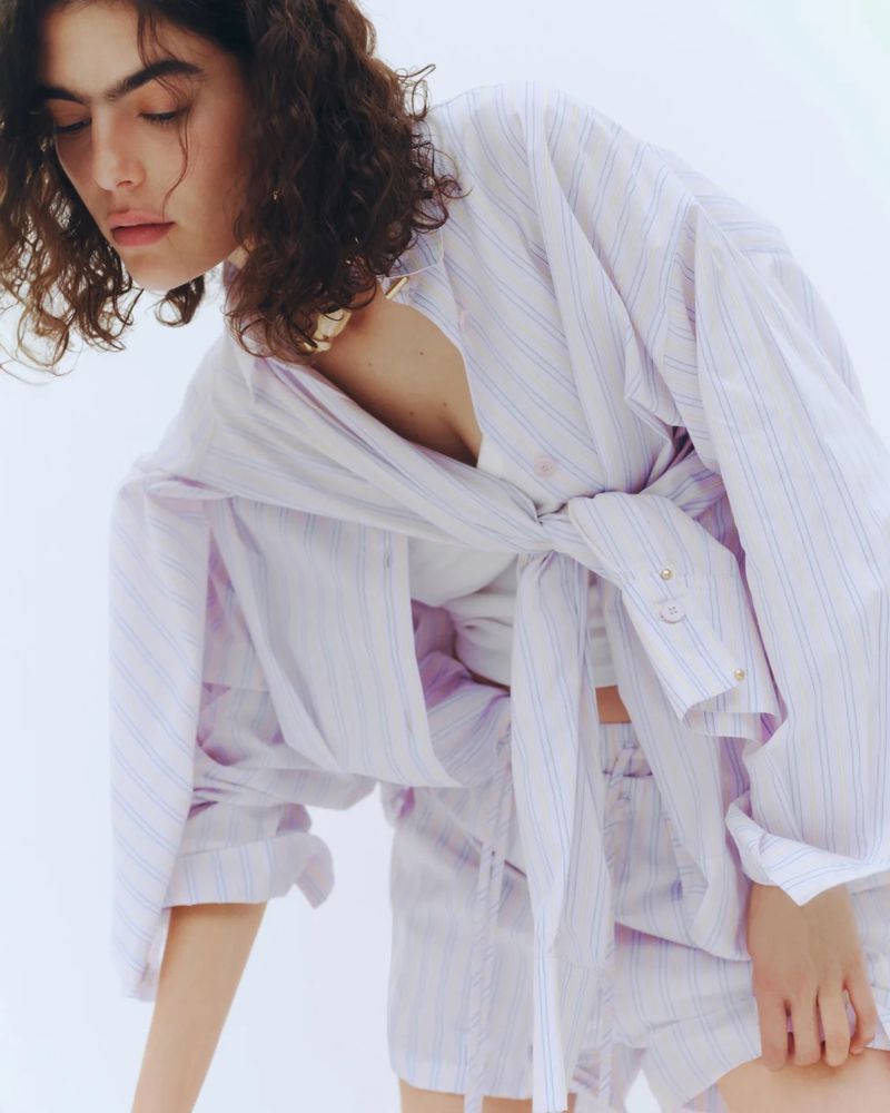Model Julia Pacha looking downward wearing a striped oversized tied shirt and matching shorts against a white backdrop.