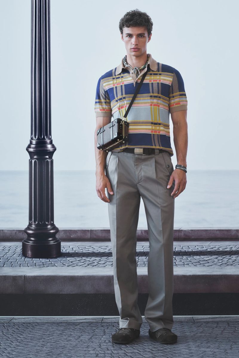 Model standing next to a black lamp post wearing a blue plaid knit polo shirt, grey trousers, and a small black crossbody trunk bag.