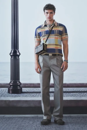Model standing next to a black lamp post wearing a blue plaid knit polo shirt, grey trousers, and a small black crossbody trunk bag.