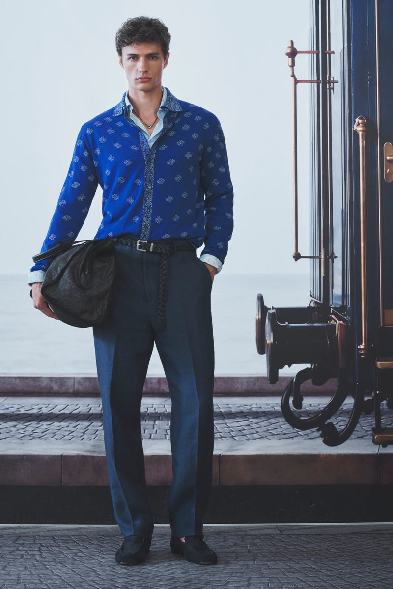 Man wearing a royal blue patterned shirt and dark trousers standing beside a vintage blue train carriage while holding a duffel bag.