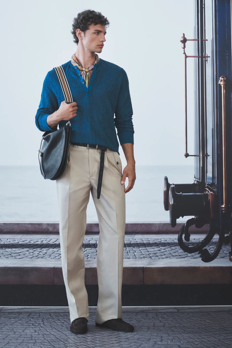 A male model wearing a teal buttoned top and cream trousers standing by a train carriage on a stone platform.