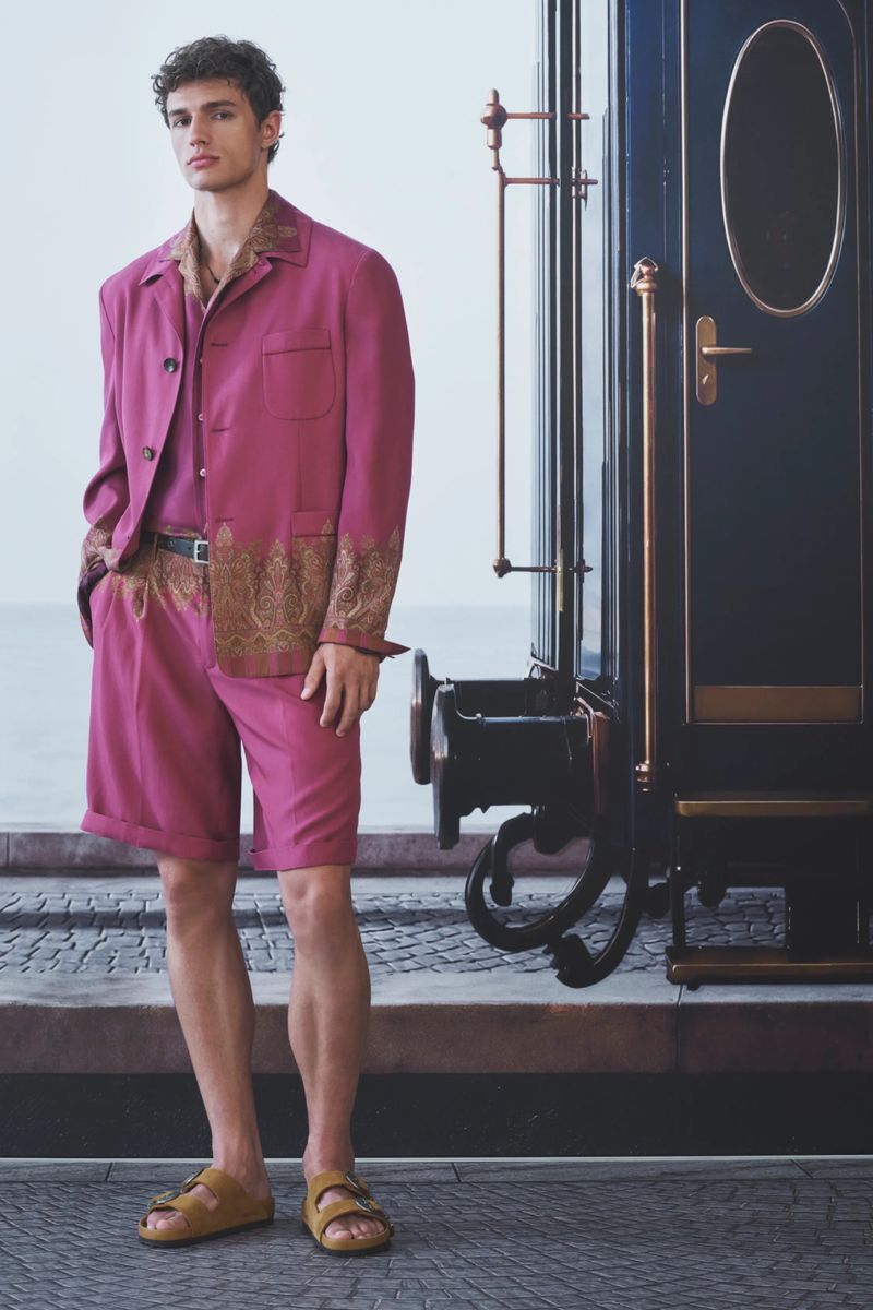 Male model in a magenta paisley-trimmed jacket and matching shorts standing beside a vintage train carriage.