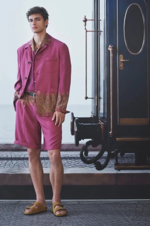Male model in a magenta paisley-trimmed jacket and matching shorts standing beside a vintage train carriage.