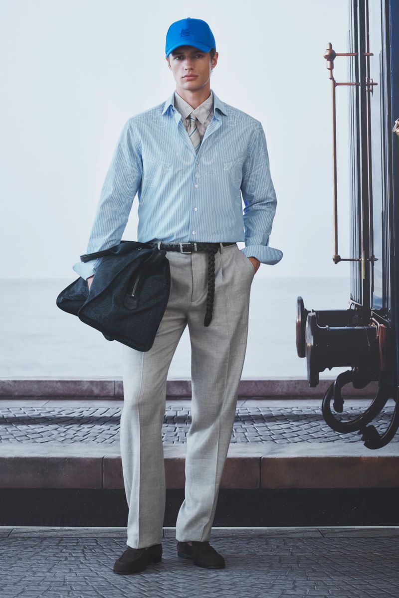 Model standing by a train car wearing a bright blue baseball cap, light blue button-down shirt, grey pleated trousers, and holding a black bag.