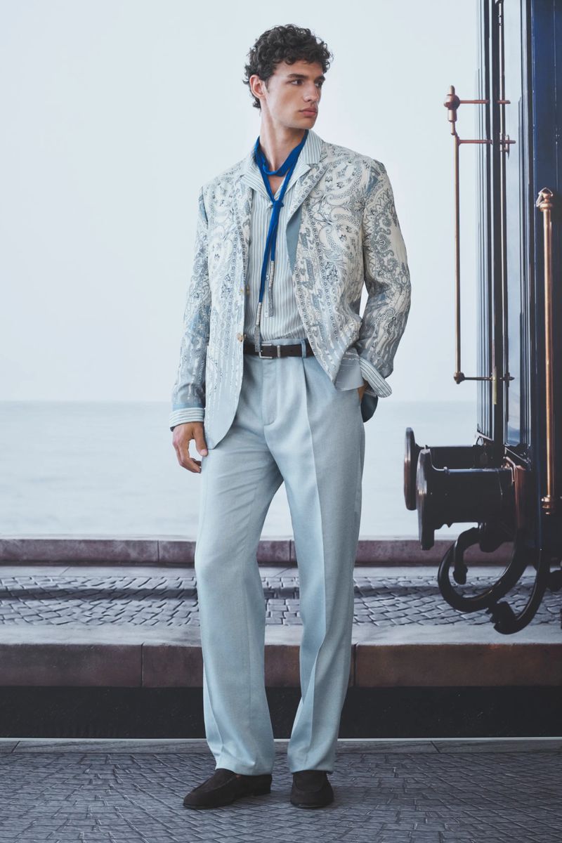 Male model in an ornate paisley blazer and light blue pleated trousers posing beside a dark train car.