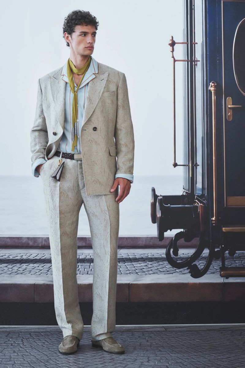 Model wearing a textured beige double-breasted suit and yellow scarf standing beside a vintage blue train carriage.
