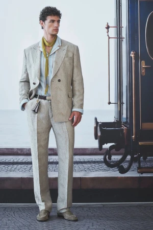 Model wearing a textured beige double-breasted suit and yellow scarf standing beside a vintage blue train carriage.