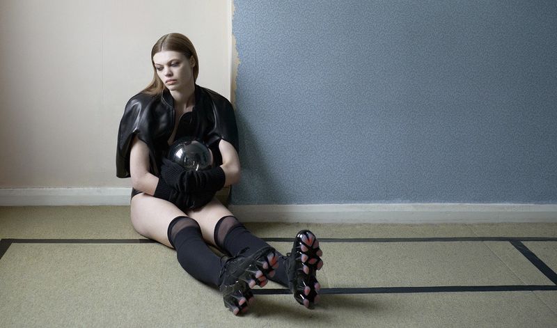 Lexie Tapper seated against an interior wall wearing black leather outerwear, knee-high socks, and futuristic sculptural sneakers.