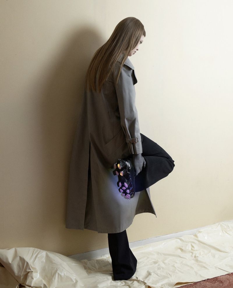 Model in grey trench coat leaning against a wall holding a black sneaker, for Nike First Sight by Karl Felix.