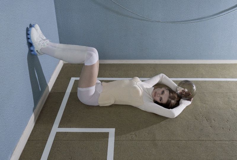 Model Lexie Tapper lying on a green court floor with legs raised against a blue wall, holding a mirrored sphere.