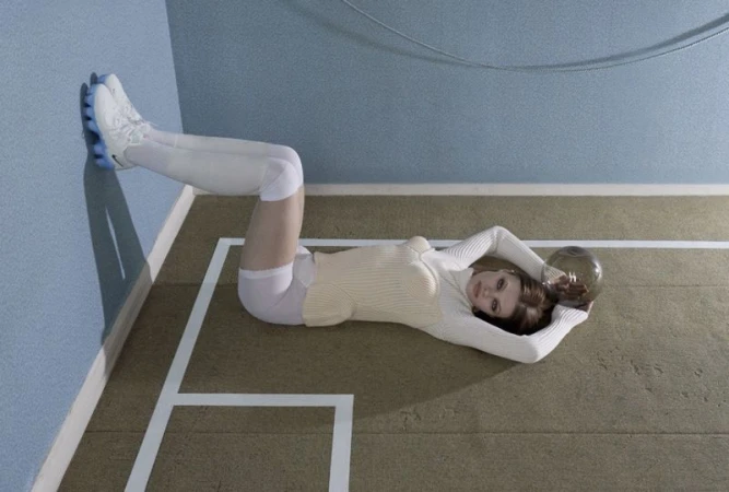 Model Lexie Tapper lying on a green court floor with legs raised against a blue wall, holding a mirrored sphere.