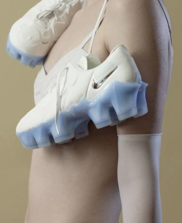 Close-up of a model wearing a sheer top with white and translucent icy blue sneakers draped across her shoulder on a beige background.