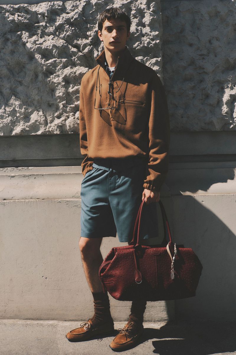 Model posing in a brown quarter-zip sweater, blue shorts, suede loafers, and holding a large burgundy leather travel bag.