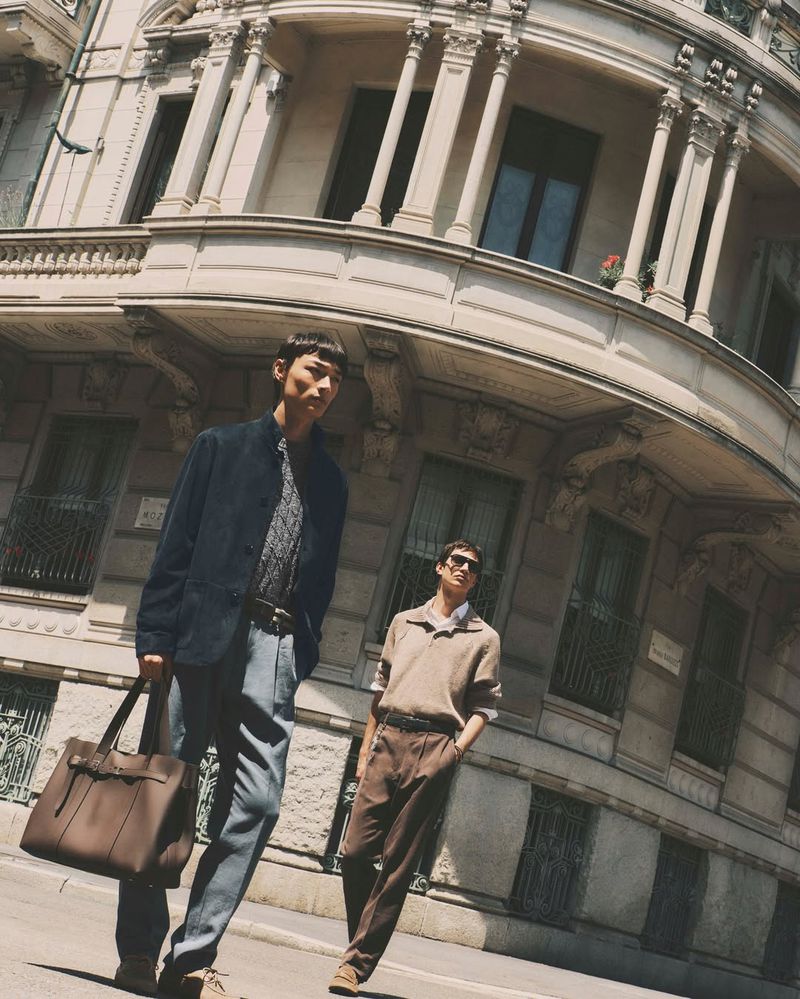 Two male models walking down a city street in front of an ornate classical building, one carrying a large brown leather tote bag.
