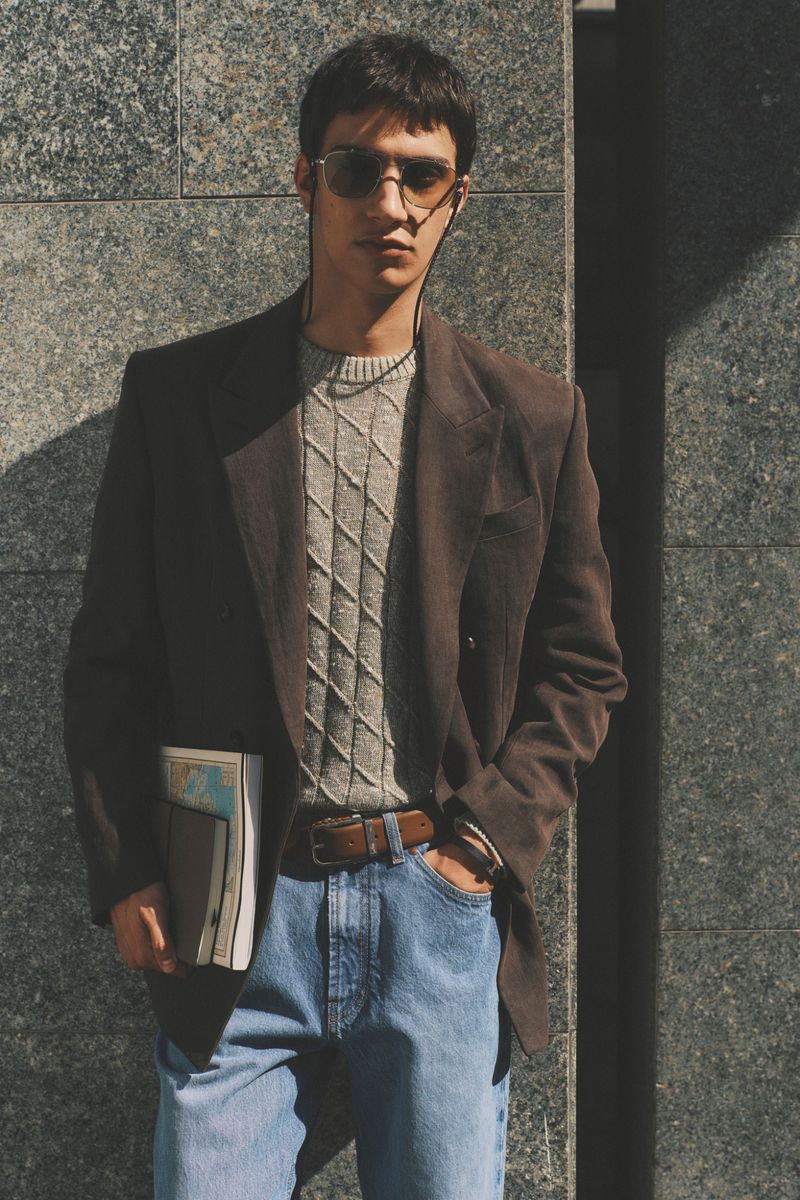 Man in a brown blazer, cable-knit sweater, tinted sunglasses, and blue jeans holding books against a stone wall.
