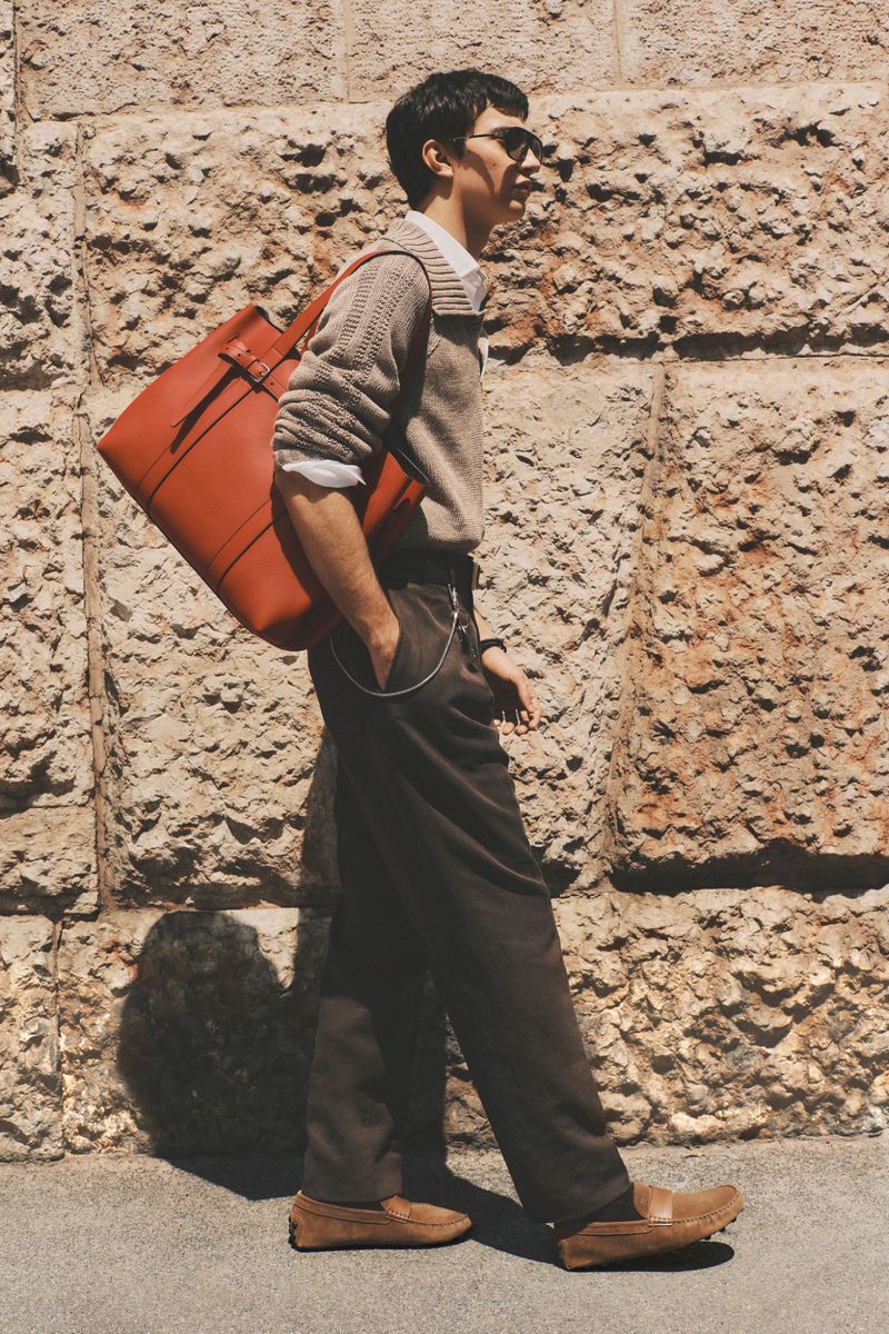 Male model in a beige knit sweater and brown trousers walking with an orange leather tote bag beside a stone wall.