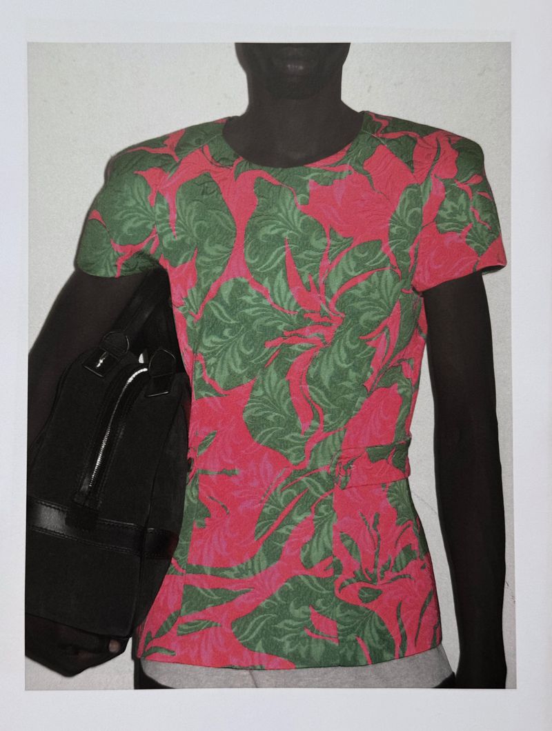 Dark-skinned man wears a pink and green floral t-shirt, carrying a black bag over his shoulder, against a plain white wall.