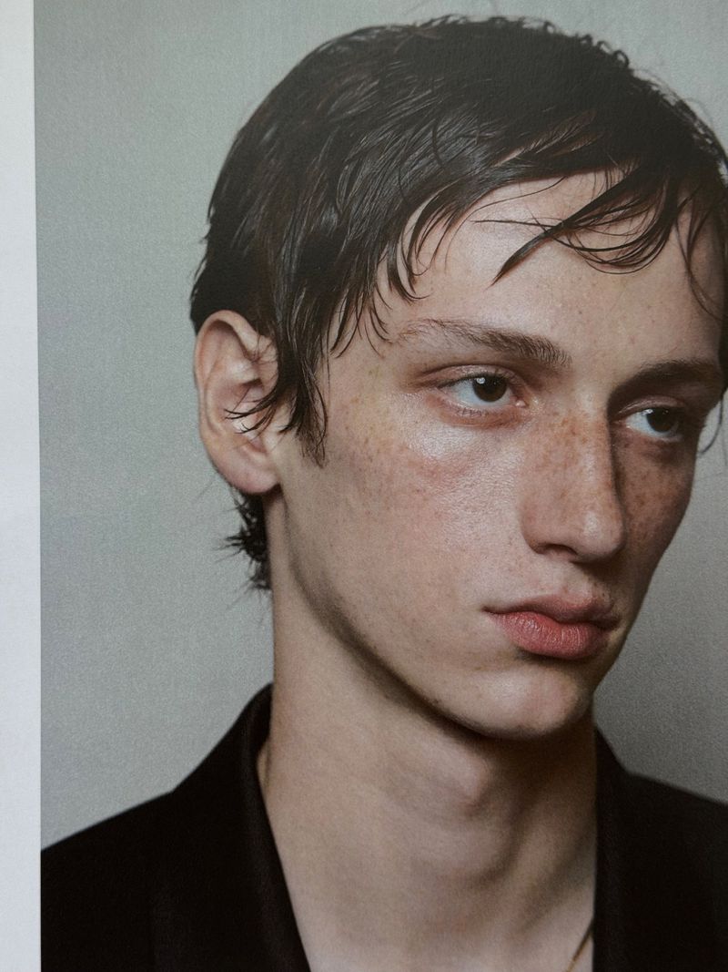 Close-up studio shot of a young adult male with dark hair, freckles, wearing a dark jacket against a neutral background in natural light.