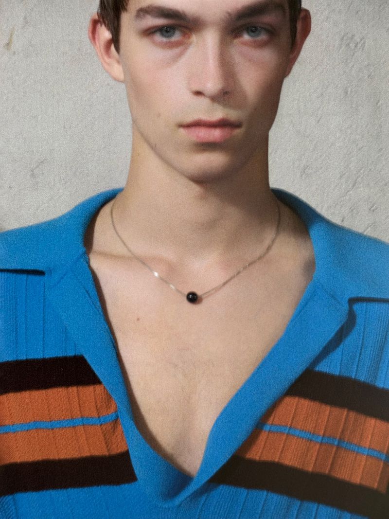 Close-up studio shot of a young adult male in a turquoise and orange striped top, wearing a necklace with a dark bead.