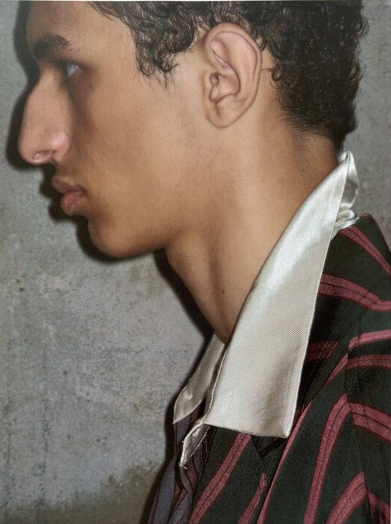 Close-up profile of a young adult male with curly hair wearing a striped shirt with a white collar against a grey concrete background.