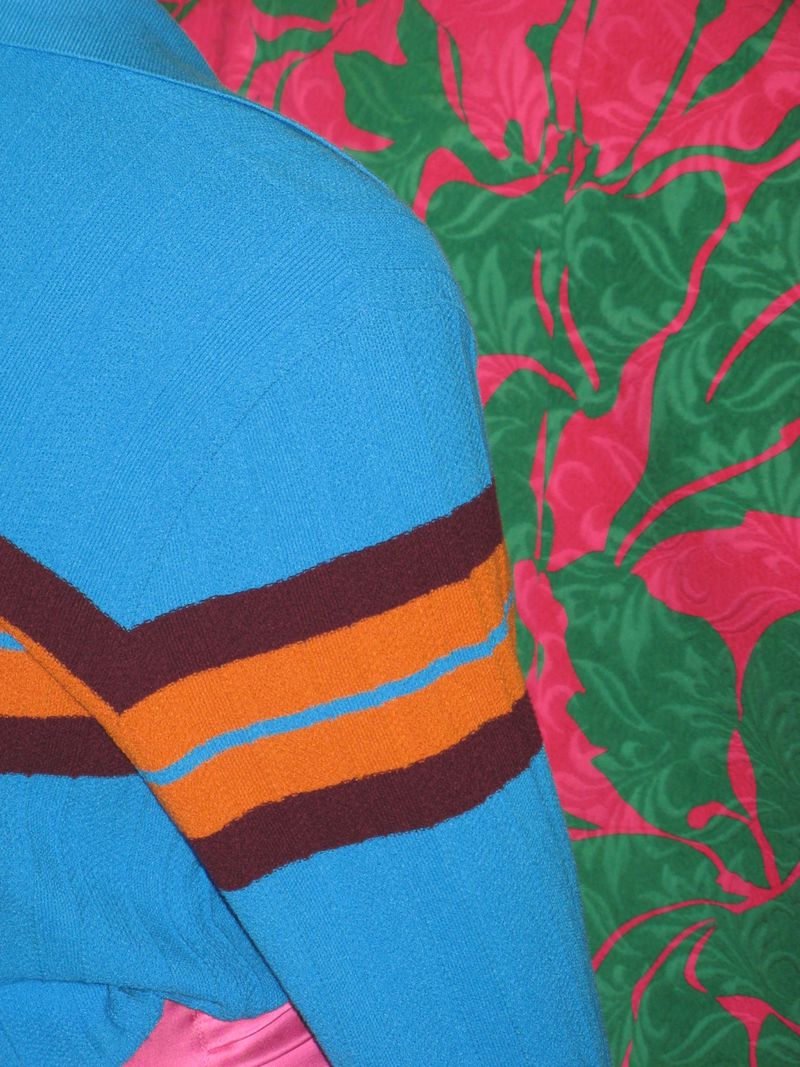 Close-up of a turquoise knit polo shirt with brown and orange stripes against a pink and green floral background, fashion apparel.