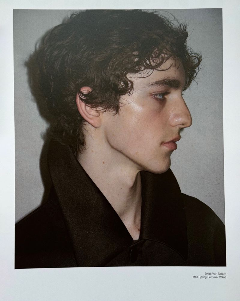 Profile shot of a young adult male with brown curly hair wearing a dark collared shirt against a neutral grey background.