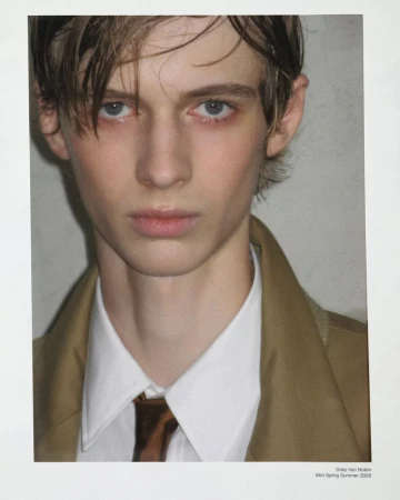 Close-up fashion portrait of a young adult male model with light skin and brown hair, wearing a beige coat, white shirt, and brown tie.