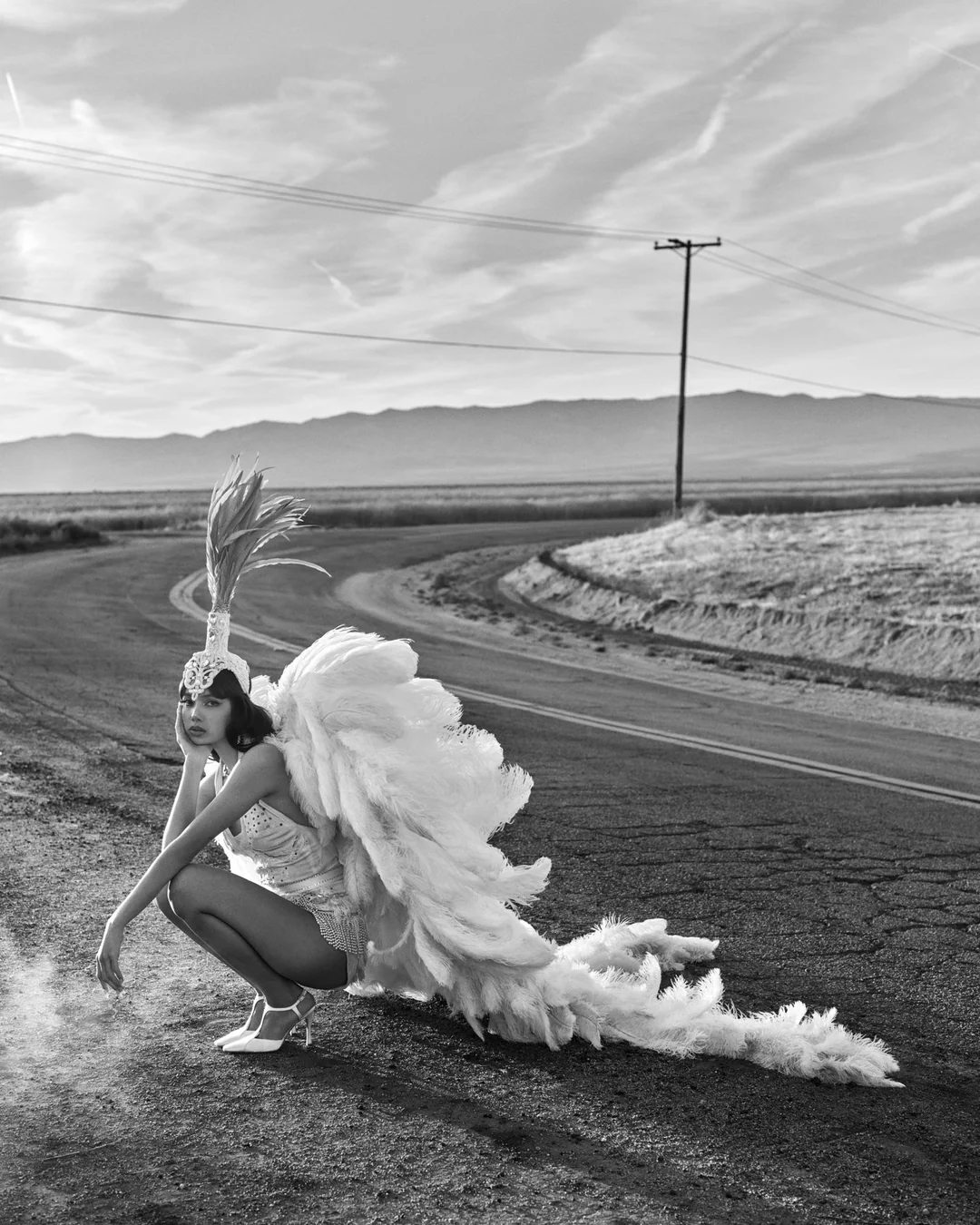 Lisa crouching on an open road wearing large white feather wings and a feathered headdress in a black and white desert editorial.