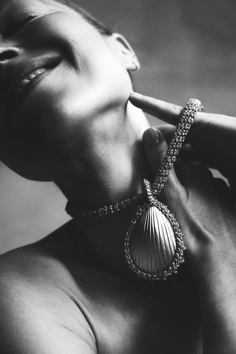 Monochrome close-up of model Heloise Guerin wearing a large ribbed teardrop pendant necklace while touching her jaw.