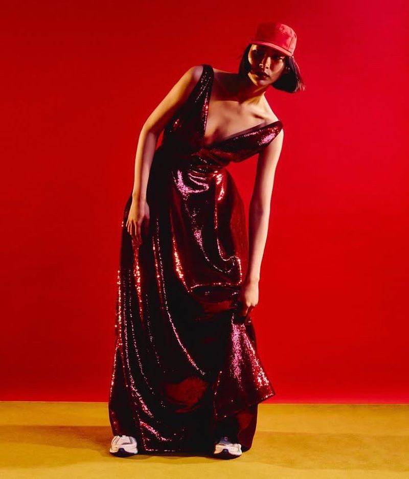 Full-length portrait of a woman in a red cap and sparkling gown, posing in a studio with a red backdrop and yellow flooring.