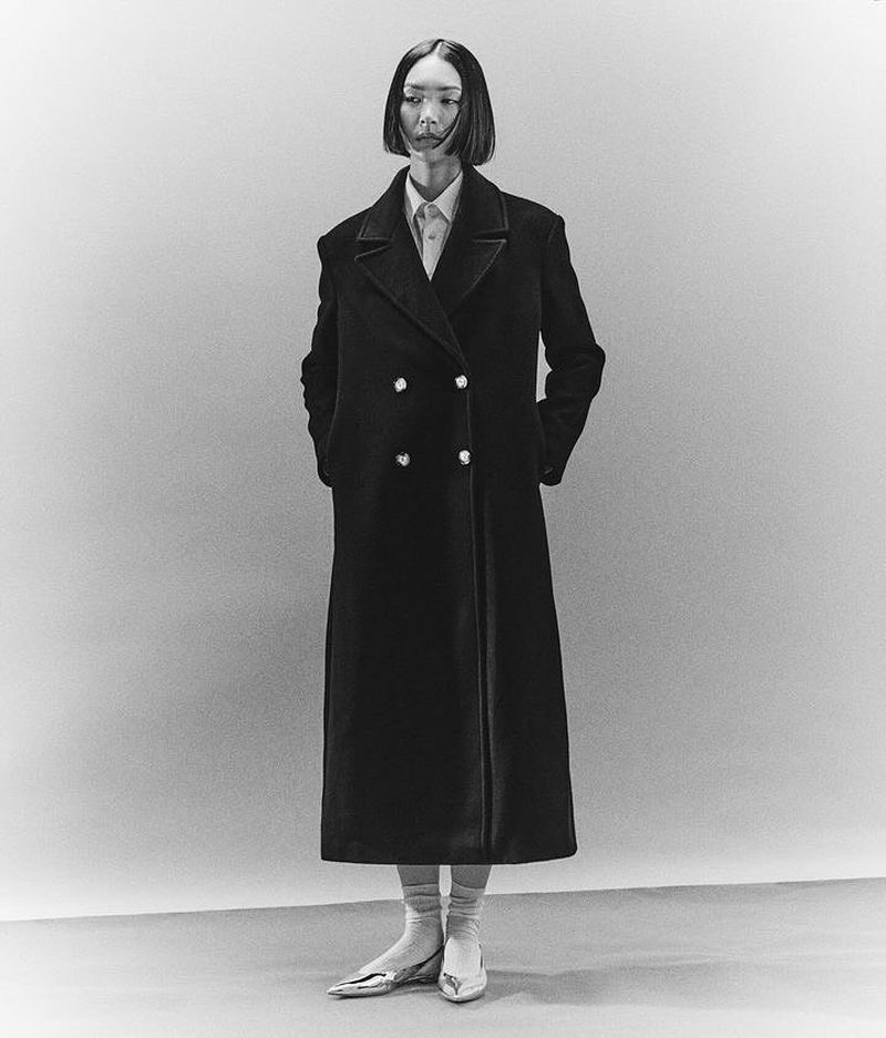 Full-length studio shot of an adult female wearing a long dark coat over a collared shirt, with metallic pointy shoes on a grey background.