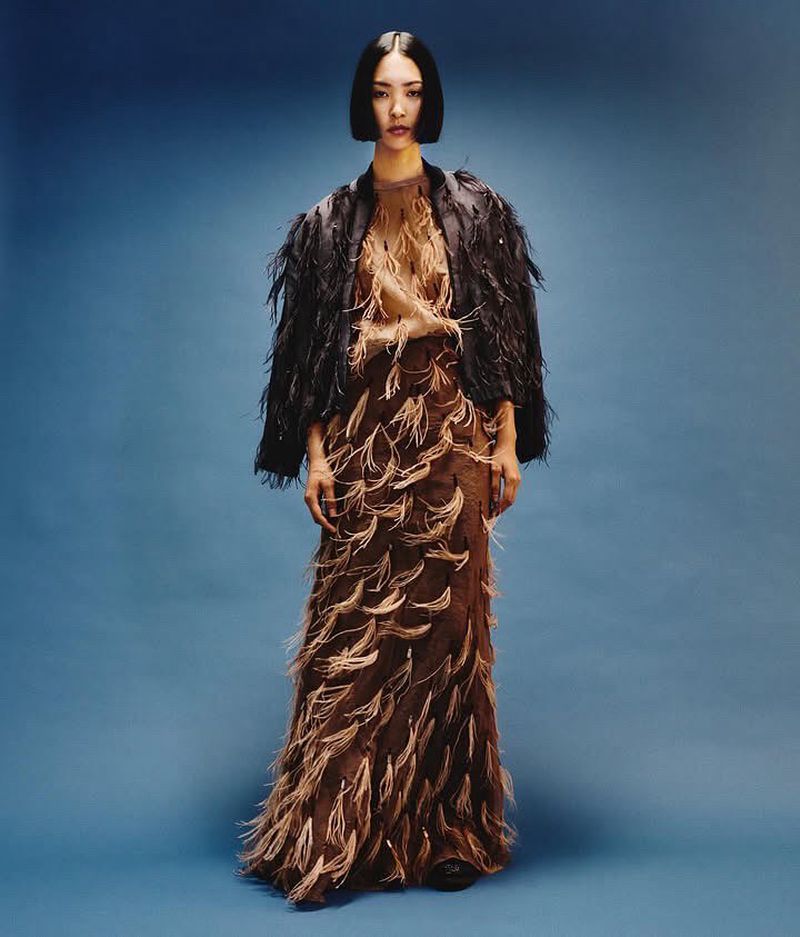 A full shot of a woman in a brown feathered dress and a black bolero jacket against a blue background in a studio setting.