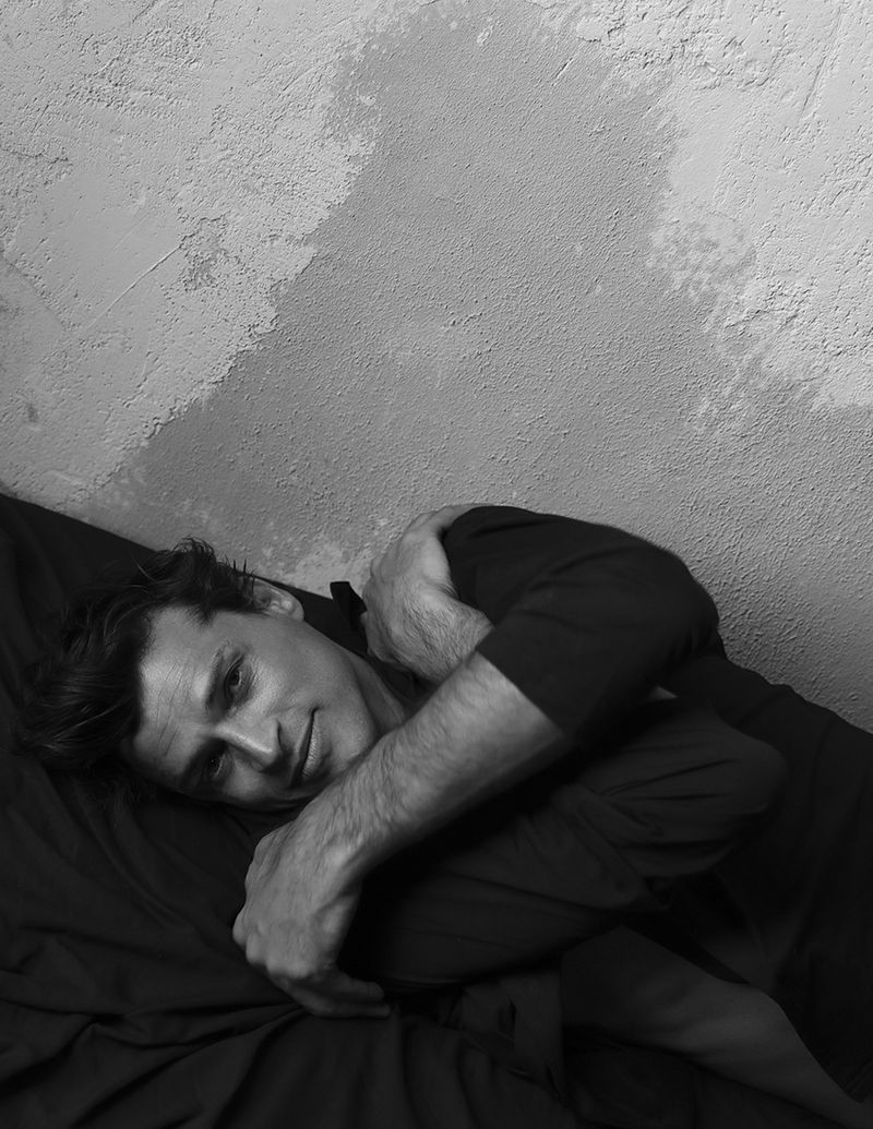 Black and white portrait of model Romulo Pires lying on dark sheets against a textured plaster wall.