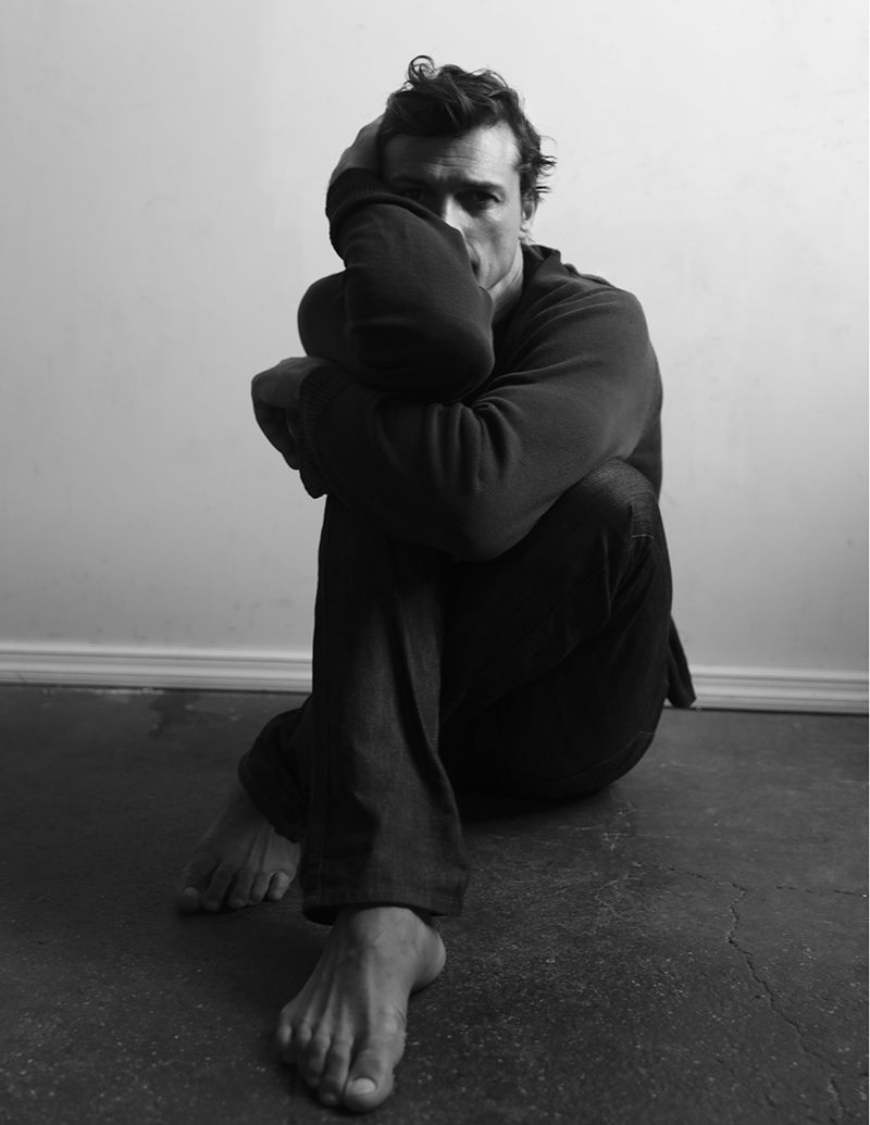 Monochrome portrait of model Romulo Pires seated barefoot on the floor with knees pulled to his chest in a dark sweatshirt.