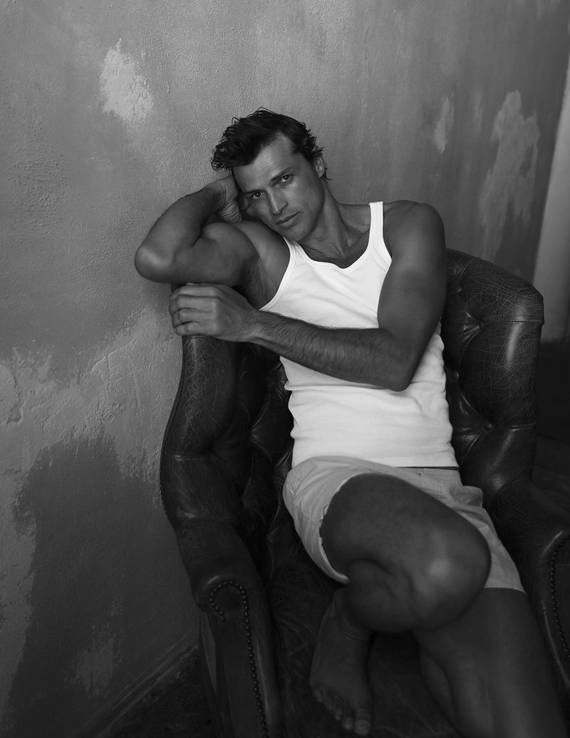 Romulo Pires seated in a tufted leather armchair wearing a white tank top against a textured wall in monochrome.